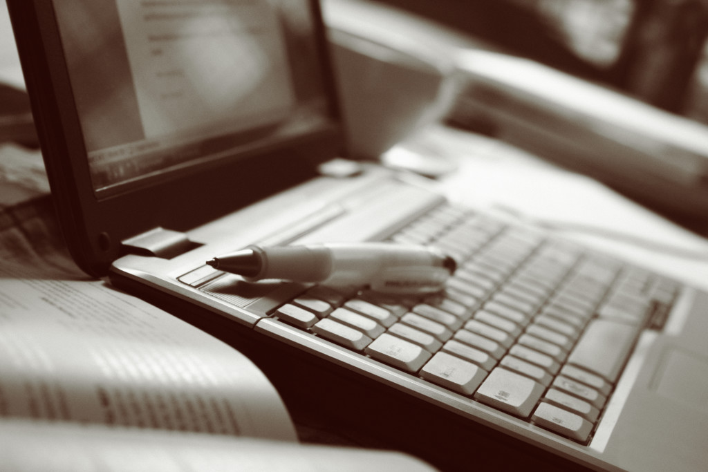 Open laptop with pen, open book and coffee cup on a table. Added grain, sepia toned image.